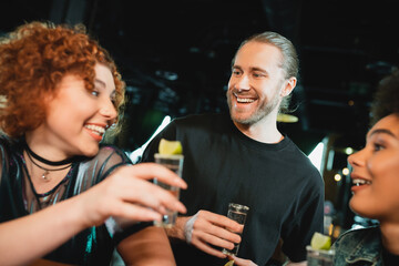 Smiling bearded man looking at blurred interracial friends with tequila in bar.