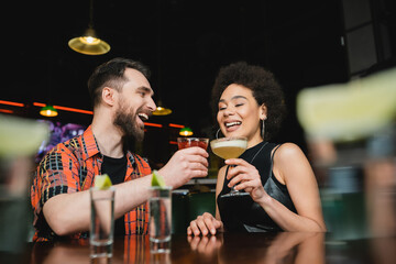 Overjoyed interracial friends holding cocktails near tequila shots on stand in bar.