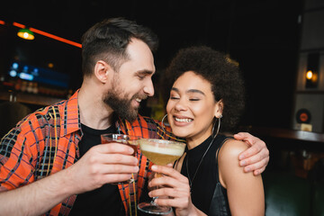 Positive man holding negroni cocktail and hugging young african american friend in bar.