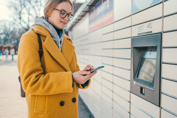 Woman holding smartphone using red automated self service post terminal machine or locker to deposit the parcel for storage. High quality photo