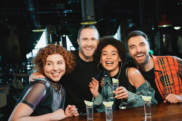 Cheerful interracial friends looking at camera near glasses with tequila in bar.