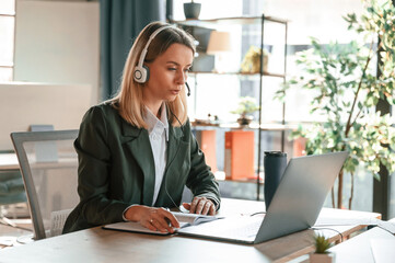 In headphones, sitting by table with laptop. Young beautiful woman in formal clothes is working in the office