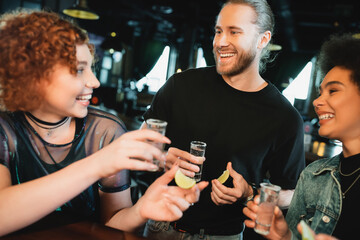 Positive multiethnic women holding tequila and lime near bearded man in bar.