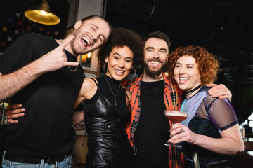 Cheerful redhead woman holding cocktail near multiethnic friends looking at camera in bar.