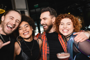 Cheerful bearded man hugging interracial friends with cocktail having fun in bar.