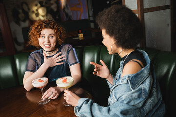 Cheerful interracial girlfriends talking near foam cocktails in bar at night.