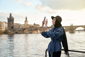 Obraz premium Stylish young woman traveller wearing black hat sitting taking pictures with her smartphone on Vltava river shore in Prague with Charles Bridge on background.
