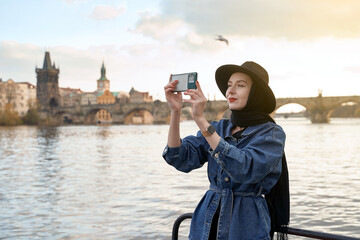Stylish young woman traveller wearing black hat sitting taking pictures with her smartphone on...