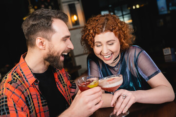 Cheerful redhead woman laughing and holding foam cocktail near bearded friend in bar.