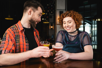 Positive man holding negroni cocktail and talking to redhead friend in bar.