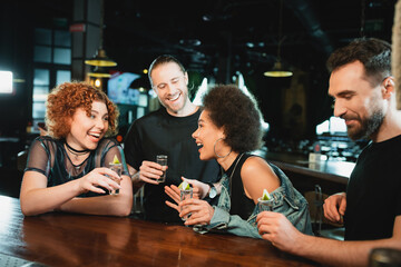 Smiling interracial women holding tequila glasses near bearded friends in bar.
