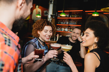 Smiling multiethnic women clinking cocktails near blurred friends in bar.