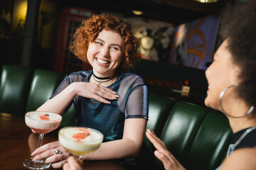 Positive redhead woman holding clover club cocktail near african american friend in bar.