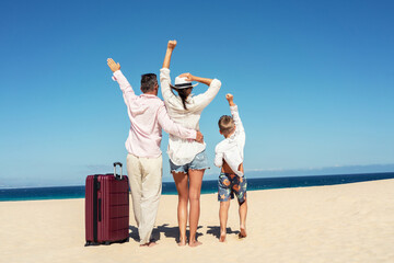 Concept of family vacation and tourism. Parents with their son standing on the beach with arms raised up, with suitcases.