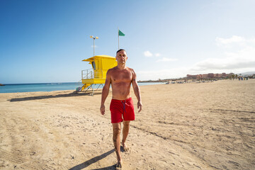 Handsome man walking relaxed on the sandy beach after swimming in the ocean. Lifeguard. Summer...