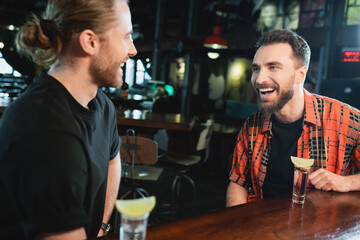 Positive bearded men talking near shots of tequila with lime in bar at night.