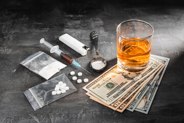 Alcohol drink in a glass, syringe with a dose of drugs, white pills in a transparent bag, narcotics powder in a spoon and US dollar cash on dark background. Concept of addiction and bad habits