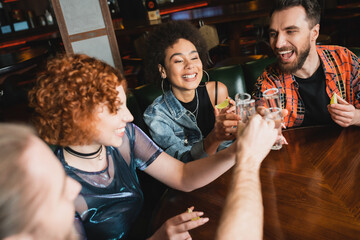 Positive interracial friends holding glasses of tequila and fresh lime in bar.
