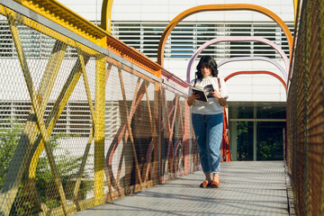 young woman walking on a pedestrian bridge reading a book