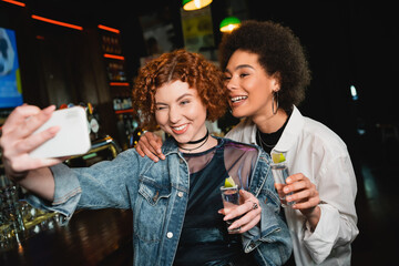 Carefree multiethnic girlfriends taking selfie and holding tequila in bar.