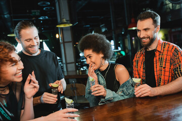 Cheerful african american woman holding glass of tequila with lime near friends in bar.