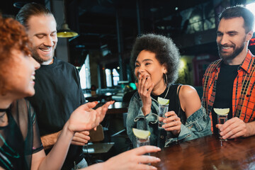 African american woman laughing near friends with tequila shots in bar.