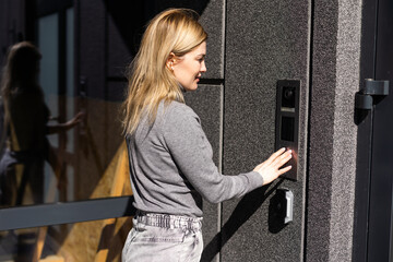 Happy woman ringing on doorbell at building entrance. Using intercom