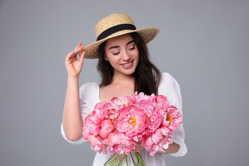 Beautiful young woman in straw hat with bouquet of pink peonies against grey background