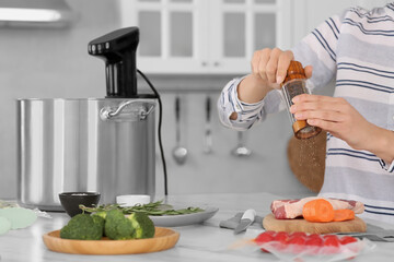 Woman grinding pepper onto meat near pot with sous vide cooker in kitchen, closeup. Thermal...