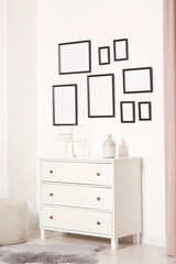 Empty frames hanging on white wall, chest of drawers and ottoman indoors