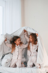 Fototapeta na wymiar Under the blanket on bed. Two little girls are playing and having fun together in domestic room