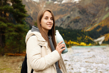 Happy young woman holding thermo bottle with drink in beautiful mountains. Space for text - obrazy, fototapety, plakaty