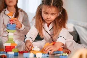 Bunch of plastic construction toys are on the floor. Two little girls are playing and having fun together in domestic room