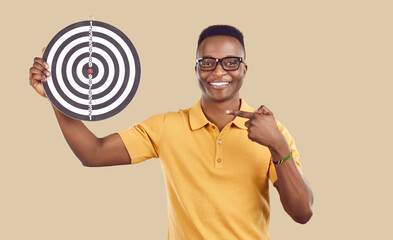 Portrait of smiling African American man isolated on brown studio background point at dartboard. Happy biracial guy show at darts board as concept of reaching goal and aim achievement.