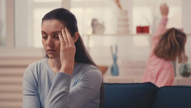 Exhausted young mother sitting on sofa while little daughter runs around, adhd