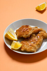 Classic fried Wiener Schnitzel served with lemon on a white plate. Breaded fried meat. A traditional dish of Austrian or German cuisine. Close-up. Copy space. Orange bright background