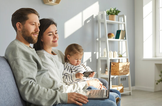 Happy family sitting on sofa and using different digital devices. Young couple watching video on laptop while their little child is playing online games on modern phone or tablet. Technology concept
