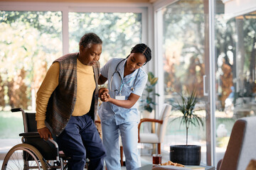 Young black nurse assists senior man in getting up from wheelchair in nursing home.