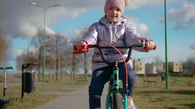 Young cyclist on thrilling ride through city park, as she pedals her bike with determination and spins wheel with enthusiasm, joyful smile in slow motion. Happy childhood, sports outdoors in
