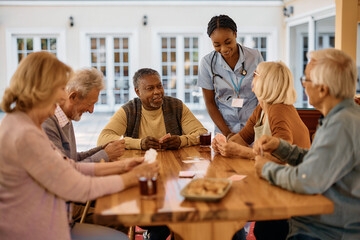 Group of happy seniors talking to young nurse while playing cards on patio in nursing home. - obrazy, fototapety, plakaty