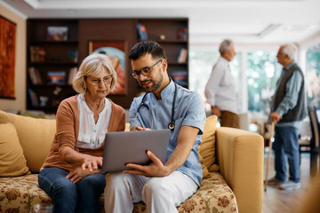 Young caregiver assists senior woman in using laptop at nursing home.