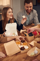 Food is in the paper eco boxes. Friends are sitting indoors at home eating macarons, desserts and blueberries together