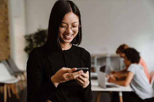 Smiling asian woman using smartphone while standing in office with her colleagues on a background