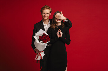 Man covering his girlfriend's eyes while giving her roses bouquet isolated over red wall