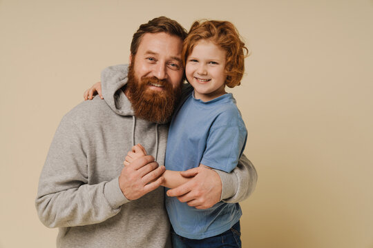 Ginger father and son smiling at camera and hugging while standing isolated over beige background