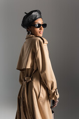 trendy african american model in beret and trench coat standing isolated on grey.