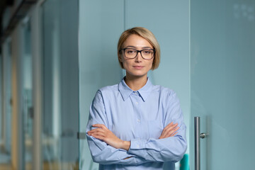 Serious and successful business woman boss inside office building at workplace, woman looking at camera thoughtfully with crossed arms and blue shirt.