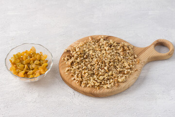 A wooden board with chopped walnuts and a bowl of golden raisins on a light gray background. Home baking ingredients