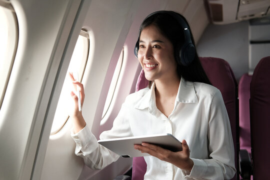 Cheerful Asian woman sitting on a plane looking out the window using a laptop and listening to music on the go