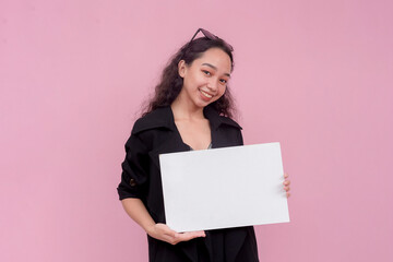 A young and pretty southeast asian lady with wavy hair holding a blank white cardboard sign. For a notice, guideline or advertisement. Isolated on a pink background.